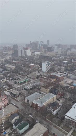 Vertical drone flying toward downtown Winnipeg Manitoba showing streets, cars, buildings, and falling snow under cloudy skies during early winter.