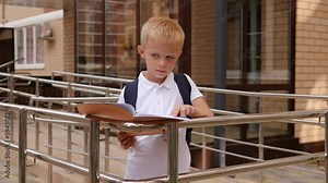 A little boy stands outside the school building and reads notes in a notebook. Elementary school.