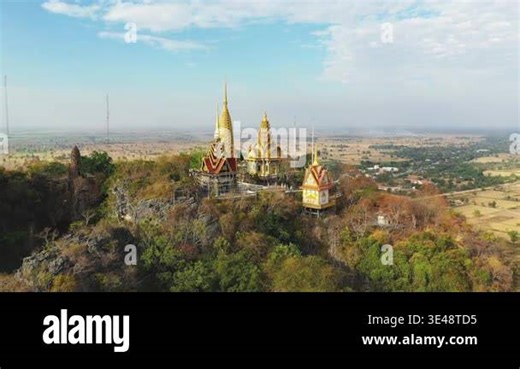 Aerial view of a golden temple complex perched atop Phnom Sampov mountain, surrounded by rural Cambodian landscape under a partly cloudy sky Stock Video Footage - Alamy