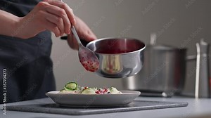 A female chef in a white uniform and a black apron in the restaurant kitchen. Cooking. The cook pours red cranberry salad sauce. Making and decorating food.