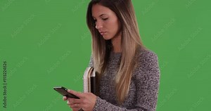 Millennial woman student texting friends on cell phone while holding books on greenscreen. Young Caucasian college student messaging study group on smartphone. 4k on green for keying or compositing