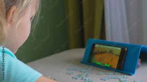 In a room with green wallpaper and yellow wallpaper, a girl with blond hair and a blue T-shirt is sitting at the table, watching a basketball game on a smartphone, next to the table are puzzle pieces