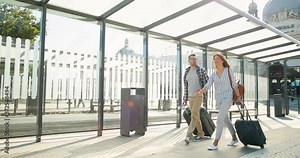 Caucasian happy young married couple in love walking at bus stop and carrying suitcases on wheels. Beautiful woman and handsome man travelling and strolling at train station.