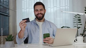 Contented male holding bank card and modern mobile phone with smile while sitting at home office. Successful male in shirt using contactless online payment for ordering shipping and looking at camera.
