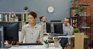 Caucasian attractive woman sitting at the computer and taping on her workday in office while her male co-workers working on the background at the office.