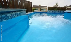 A rectangular swimming pool with concrete edges is located in the backyard of the house