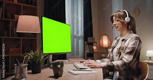 Female sitting in front of chroma key computer. Woman using headphones for online meeting. Female looking at mock up green screen. Woman conducting online conference. Female and chroma key template.