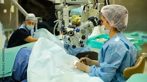 Female Doctor Conducting Eye Surgery Under the Microscope in the Operating Room and Other Doctor Watching Her. Ophthalmology. Sight Restoration