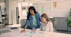 Two workmates review paperwork with infographics include various types of color charts and graphs, learn sales in visual data, discuss marketing trends, make statistics analysis, engaged in teamwork