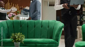 Focused hotel manager working on laptop sitting in lobby, looking at screen and keeping track of reservations to manage bookings. Bellboy in uniform works in hospitality industry. Handheld shot.