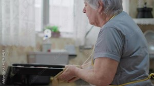 Candid Domestic Elderly Lifestyle: Senior Woman Taking Food Out of the Oven, Older Lady Standing in Kitchen, Wearing Apron and Cooking Meal
