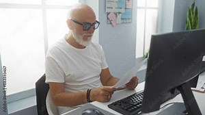 Man reading documents at a desk in a modern office with a computer screen and paperwork, showcasing a senior professional engaged in office work under natural light from large windows