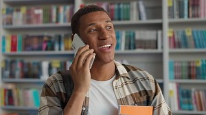 Happy African American man talking mobile phone smartphone technology communicate conversation library student learner university chatting smiling male guy bookshelf educational study calling laughter