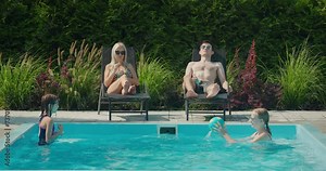 The family relaxes by their pool in the backyard of the house. Adults sunbathe on sun loungers, two children swim in the pool