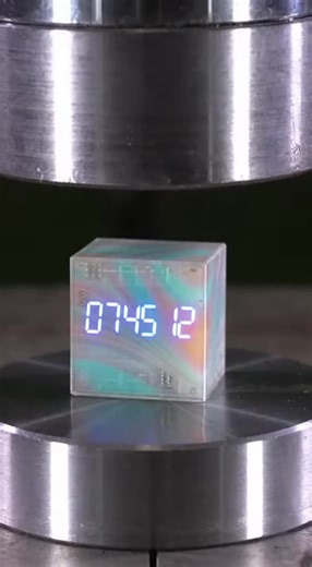 Watch as a stunning opal-tinted glass digital alarm cube meets the hydraulic press. The LED digits flicker and freeze mid-count as the press descends, then the entire cube cracks and shatters into sharp fragments with minimal pearlescent seep.
