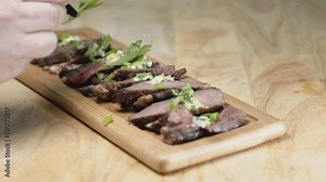 Slow Motion Slider Shot of Medium Rare Sliced Steak on a Rectangular wooden Chopping Board With Food Stylist Hands Adding Garnish