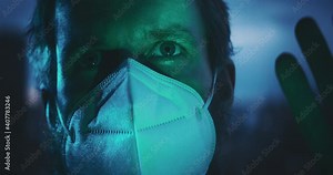 Man in a face mask placing his hand on a window and looking out