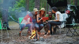 Company of five amazing friends hanging out in the woods sharing tablet computer screen watching movies near campfire. Technology and nature. Modern camping.