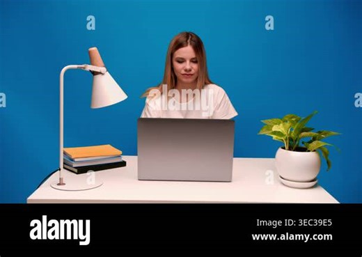 Beautiful young female student or freelancer sitting at a white desk, typing on her laptop and smiling in front of a blue background Stock Video Footage - Alamy
