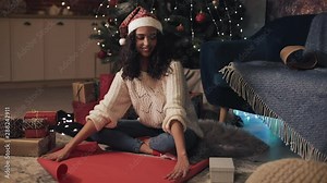 Young Beautiful Girl in Santas Hat Sitting under Decorated Cristmas Tree Deploying the Scroll of Paper, Putting Present Box on it. Holiday Celebration Concept.