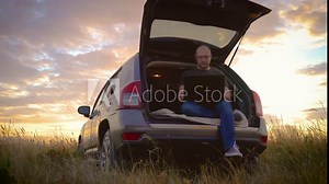 Concentrated freelancer man working remotely online typing internet communication technology sitting in car trunk while traveling, general plan in field at sunset. Busy worker freelancer entrepreneur