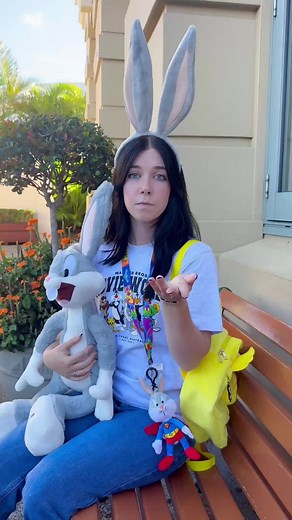 Repeat offender: putting my Premium Perks to work. 😏 Did you know Premium Annual Passholders now get 20% off retail shopping in-park? 🛍️ T&Cs, exclusions apply. ID: A woman wearing oversized bunny ears sits on a bench holding a Bugs Bunny plush toy while showing off Movie World merchandise including a graphic T-shirt and yellow backpack.