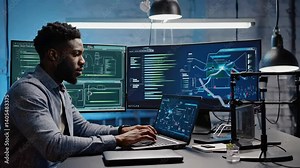 Focused developer coding on laptop in modern office with multiple monitors displaying complex graphs and data analysis tools.