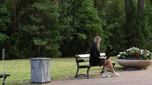 Businesswoman sitting on bench in summer park using, typing on laptop computer, studying outdoors. Female blond person working remote.