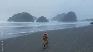 Aerial back view traveler woman walking by wet ocean beach leaving footprint on sand surface. Oregon coast beach on cloudy day with black rocks on background. Backpacker tourist enjoying nature 4K