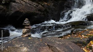 Stationary low view at stack of rocks cairn on edge of cascading river with fall leaves