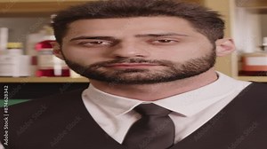 Vertical Tilt up medium portrait of young brutal Middle Eastern restaurant bartender in uniform standing against shelves with expensive alcohol and looking at camera