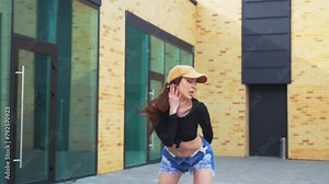 Woman in denim shorts and black top gracefully dancing in high heels in front of urban building. Slow motion shooting. Energetic woman performing dance routine outdoors. Dancer expressing joy in city