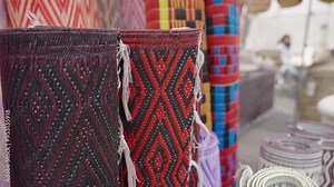 A close-up view of intricately woven pattern rugs displayed in a bustling street market. The rich colors and textures highlight the craftsmanship and local artistry.