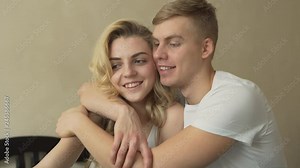 Beautiful blond couple/lovers dressed in white sitting on a bed and hugging/cuddling each other while taking photos, smiling and posing to a camera