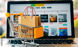 A miniature shopping cart with a brown paper bag inside, sits in front of a laptop computer screen with an online store displayed.