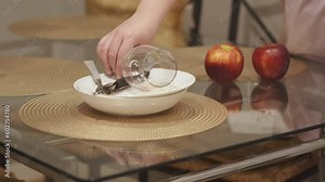 Housewife stacks dirty dishes after eating on table with round golden placemats. Unrecognizable woman puts wine glass on plate with cutlery. Two apples lie on dirty table.
