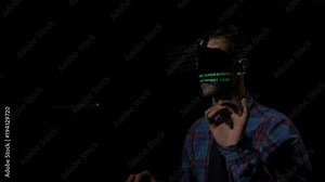 Young male hacker with VR glasses cracking codes using computer from his dark room while the green data is reflected on his face