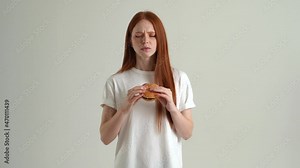Portrait of hungry young woman eating burger and suffering stomach ache, feeling nausea, food poisoning symptom, gastritis standing on white isolated background in studio. Shooting in slow motion.