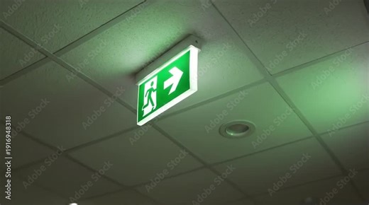 Bright Green Emergency Exit Sign Illuminating a White Grid Ceiling, Guiding to Safety with Arrow and Running Person Symbol in a Dimly Lit Indoor En...