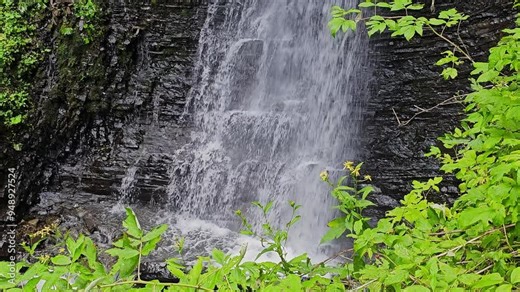 Waterfall Flowing Over Rocks with Green Foliage, A beautiful waterfall cascading over dark rocks, surrounded by lush green foliage. The water flows down in a powerful stream