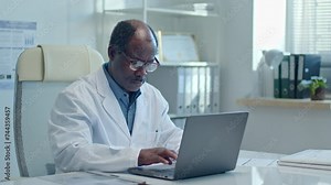 Senior African American doctor sitting at desk in medical office, opening laptop and typing on it during workday