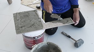 Position a piece of ceramic tile on a bucket, a construction worker is plastering the mortar to the tile as he prepares to put it in place on the floor | Premium Stock Video Footage