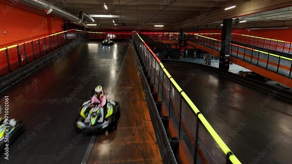 Adults in go-karts speeding around a curve on an indoor track with bright orange barriers. Go-Kart Racers Navigating Indoor Track Curve