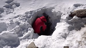A mountaineer with a backpack descends into an ice cave. There are big rocks in the hole, white snow and ice all around. A guy in a red suit explores the entrance. Trying to get inside. Mountains