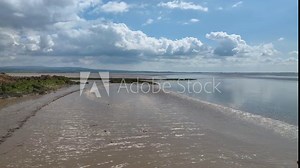 Lateral low flight along muddy beach with fluffy clouds on horizon on bright spring day at Jenny Brown's Point, Silverdale, Lancashire, England, UK.