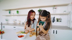 Two women are joyfully cooking together in a modern kitchen filled with fresh ingredients, showcasing their culinary skills and celebrating the joy. While daughter plays on smartphone