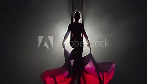 Silhouette of an aerial circus gymnast performing on aerial silk and waving canvas like wings. Beautiful circus show performed by young woman acrobat on black background with backlight. Slow motion.