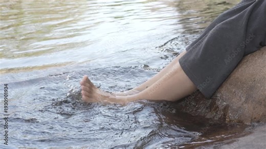 Bare feet and lower legs splashing in shallow river water beside a rock, relaxing outdoors in nature.
