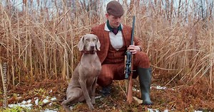 elegant hunter man sitting with dog behind grass pointing and giving instructions to dog while hunting in nature Slow motion, guy has found some game, bird or duck to shoot preparing to kill animal