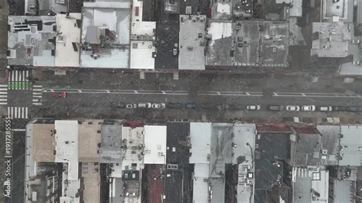 Snowy American city street during winter storm, showing dense rowhouses, parked cars and light traffic moving through an urban residential neighborhood. Aerial top down shot.